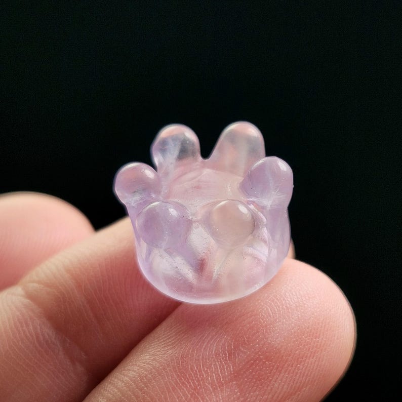 May include: A small, translucent, light purple crystal carving in the shape of a flower or hand. The object has rounded petals or fingers and a smooth, polished surface. The background is a solid black.