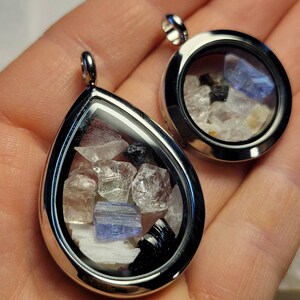 Puede incluir: Dos colgantes plateados, uno en forma de l&aacute;grima y otro redondo, cada uno con una colecci&oacute;n de piedras preciosas en bruto y coloridas. Las gemas, incluyendo piedras claras, azules y negras, son visibles a trav&eacute;s del cristal. Los colgantes tienen un acabado pulido.