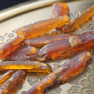 May include: Close-up of several amber-colored, translucent, elongated crystals. The crystals vary in size and shape, with some appearing more rectangular and others more rounded. They are arranged on a gold-colored, ornate tray.