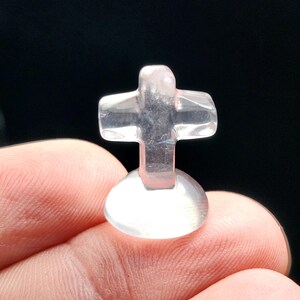 May include: A small, clear, cross-shaped crystal figurine. The cross has a rounded base and is held between fingers against a black background. The crystal is translucent with a slight pink hue, and the cross shape is clearly defined.