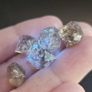 May include: A collection of raw, uncut Herkimer diamonds. These clear, double-terminated quartz crystals have a natural, faceted appearance. The crystals vary in size and clarity, with some displaying inclusions and a smoky hue. The diamonds are held in a hand.