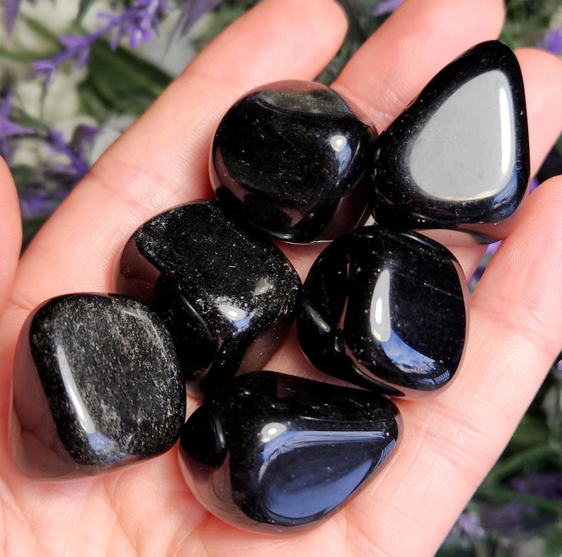 May include: A collection of six polished black stones with smooth, rounded surfaces and reflective highlights. The stones are dark, opaque, and have irregular, organic shapes, showcasing a glossy finish that catches the light across their surfaces.