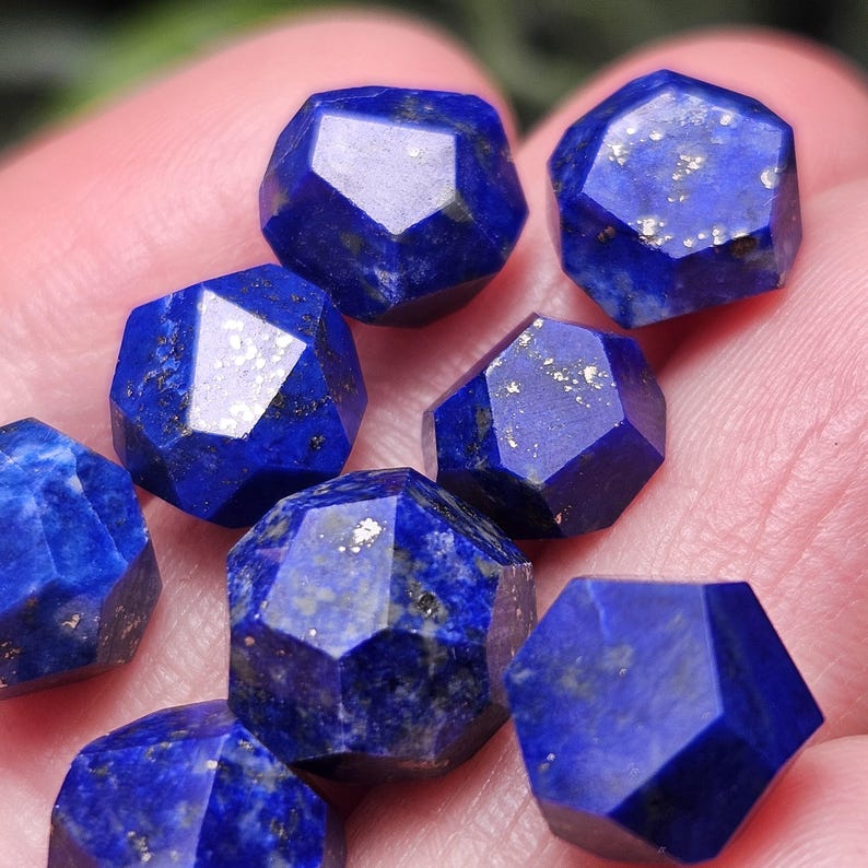 May include: A collection of seven faceted lapis lazuli gemstones, each displaying a deep blue color with natural golden pyrite inclusions. The stones are cut into geometric shapes with multiple flat surfaces that reflect light.