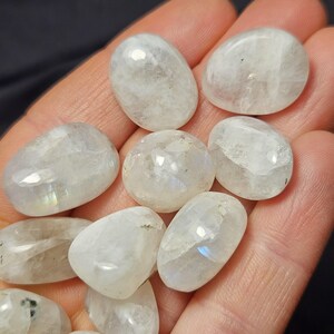 May include: A selection of polished, oval moonstone gemstones. The stones are a translucent white with subtle iridescent flashes. The gemstones are smooth and appear to be held in a hand.