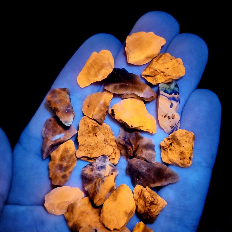 May include: A hand holding a collection of rough, irregularly shaped rocks. The rocks display a range of colors, including shades of orange, brown, and hints of blue and green. The image is illuminated with a blue light, creating a glowing effect.