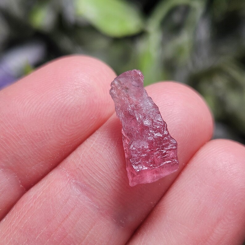 May include: A small, raw, translucent pink tourmaline crystal with a jagged, uneven surface texture and natural crystalline facets. The stone displays varying shades of deep magenta and light pink, showcasing a rough, unpolished, and organic form.