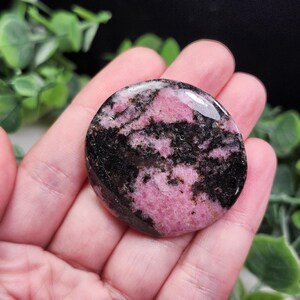 May include: A polished, circular rhodonite stone featuring a smooth, glossy surface with a distinct pattern of vibrant pink and deep black mineral inclusions. The stone is flat and round, showcasing natural color variations throughout its surface.