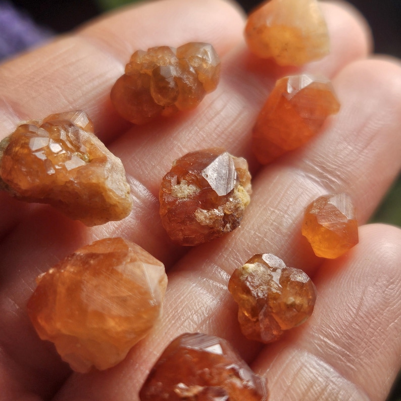 May include: A collection of raw, orange-brown gemstones, varying in size and shape, are held in a hand. The stones have a crystalline structure and a warm, earthy tone. The gemstones are likely used for jewelry or decorative purposes.