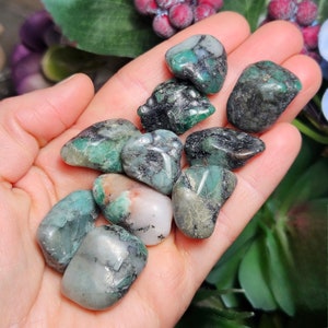 May include: A collection of raw emerald stones in various shapes and sizes. The stones exhibit a deep green hue with dark inclusions and some lighter, translucent patches. The gemstones are held in an open hand.