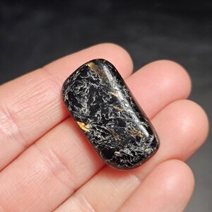 May include: A polished, rectangular black gemstone with white and gold veining. The stone is smooth and rounded, held in a hand against a dark background. The gemstone's surface reflects light, highlighting its unique patterns.