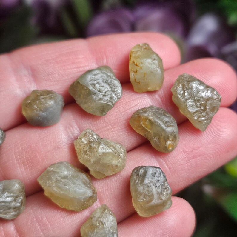 Puede incluir: Una colecci&oacute;n de gemas en bruto, sin cortar, en tonos amarillo p&aacute;lido y gris. Las piedras tienen una superficie rugosa y texturizada, con formas irregulares. Las gemas se muestran sobre un fondo borroso.