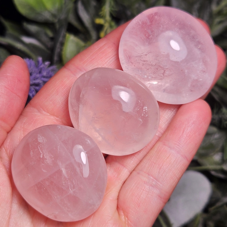 K&ouml;nnte beinhalten: Drei polierte, durchscheinende Rosenquarzsteine mit einer glatten, abgerundeten, kieselartigen Form. Die Steine weisen einen sanften, blassrosa Farbton und eine gl&auml;nzende, reflektierende Oberfl&auml;che auf, die das Licht einf&auml;ngt.