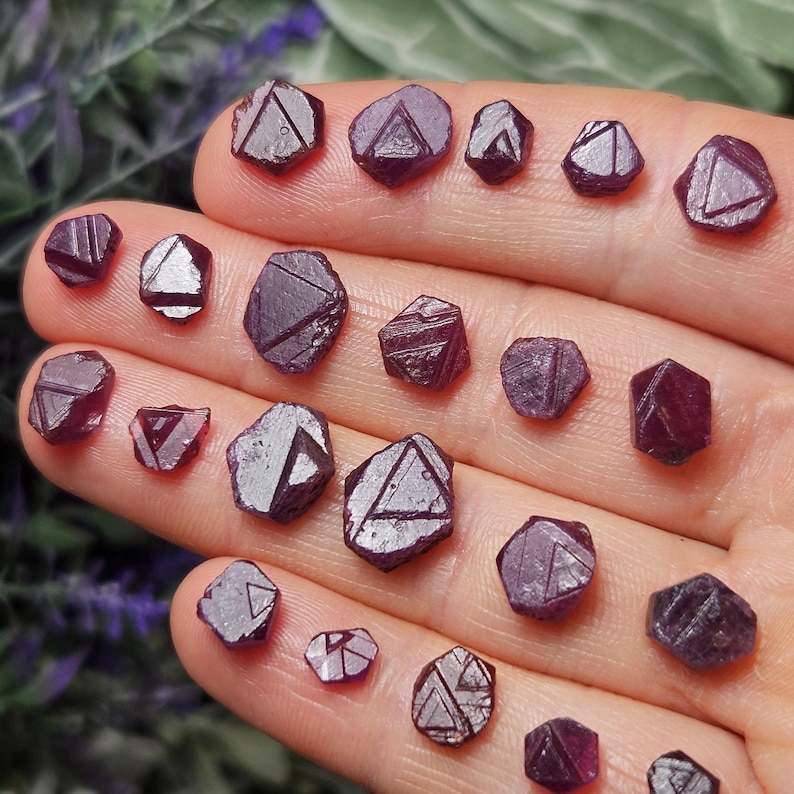 K&ouml;nnte beinhalten: Eine Sammlung von 20 kleinen, facettierten, violetten Rubinkristallen. Die Kristalle sind in verschiedenen Gr&ouml;&szlig;en und Formen, einige haben eine eher dreieckige Form, andere eine eher abgerundete Form.