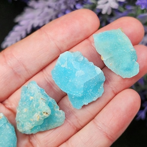 May include: Several pieces of raw, light blue aquamarine crystals are held in a hand. The crystals have a rough, textured surface and varying shapes. The background is blurred, with hints of purple flowers.