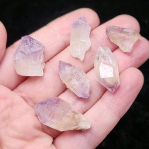 Puede incluir: Seis puntas de cristal de amatista en bruto, con un degradado de color de p&uacute;rpura a transparente. Los cristales se sostienen en una mano, mostrando sus formas naturales e irregulares y su calidad transl&uacute;cida.