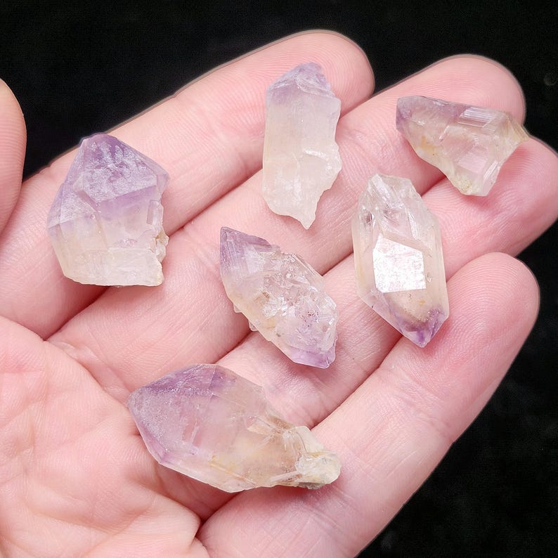 Puede incluir: Seis puntas de cristal de amatista en bruto, con un degradado de color de p&uacute;rpura a transparente. Los cristales se sostienen en una mano, mostrando sus formas naturales e irregulares y su calidad transl&uacute;cida.