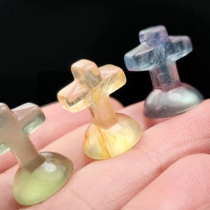 May include: Three small, translucent cross-shaped sculptures in shades of yellow, blue, and green. Each cross sits atop a rounded base. The crosses are held in a person's hand against a dark background.