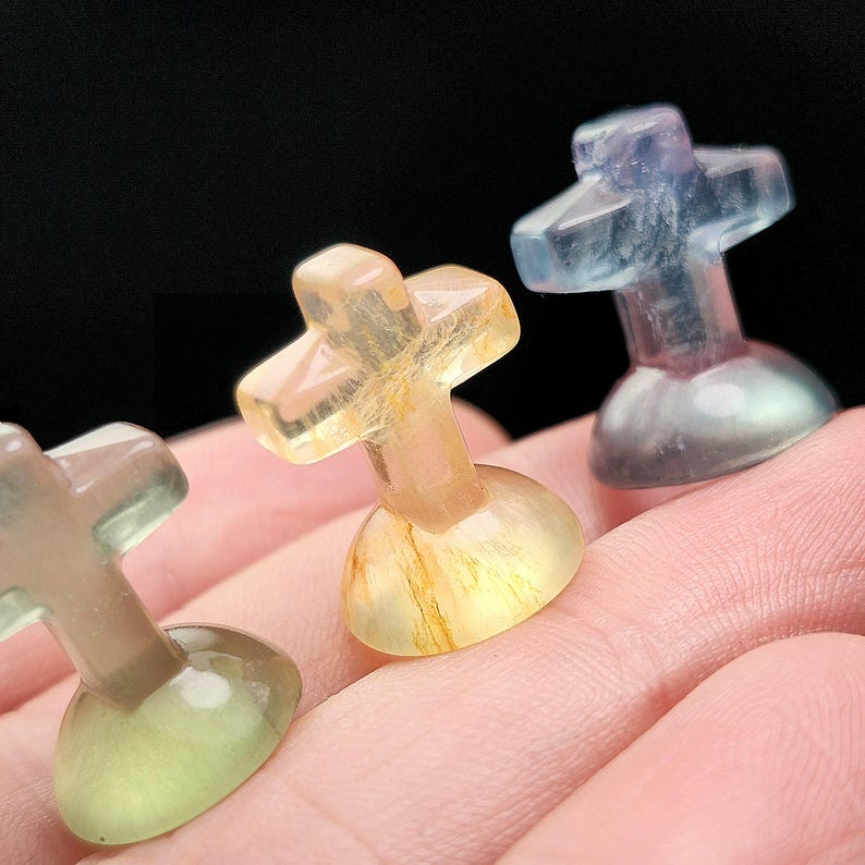 May include: Three small, translucent cross-shaped sculptures in shades of yellow, blue, and green. Each cross sits atop a rounded base. The crosses are held in a person's hand against a dark background.