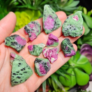 Pu&ograve; includere: Una collezione di pietre grezze di rubino in zoisite. Le pietre sono un mix di verde, nero e rosa. Il rubino rosa &egrave; visibile al centro delle pietre. Le pietre sono tenute in una mano.