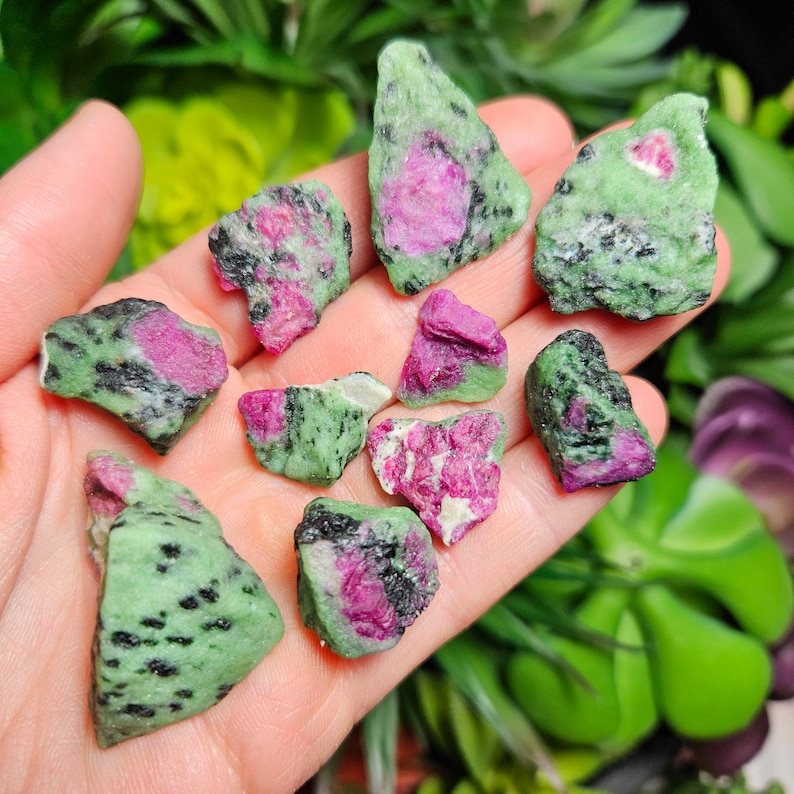 Pu&ograve; includere: Una collezione di pietre grezze di rubino in zoisite. Le pietre sono un mix di verde, nero e rosa. Il rubino rosa &egrave; visibile al centro delle pietre. Le pietre sono tenute in una mano.