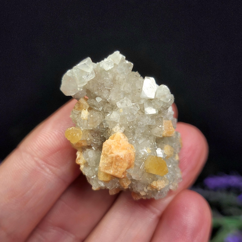 May include: A cluster of crystals in shades of gray, white, and orange. The crystals have a rough, textured appearance, with some larger, more defined points. The overall shape is irregular and organic, with a natural, earthy aesthetic.