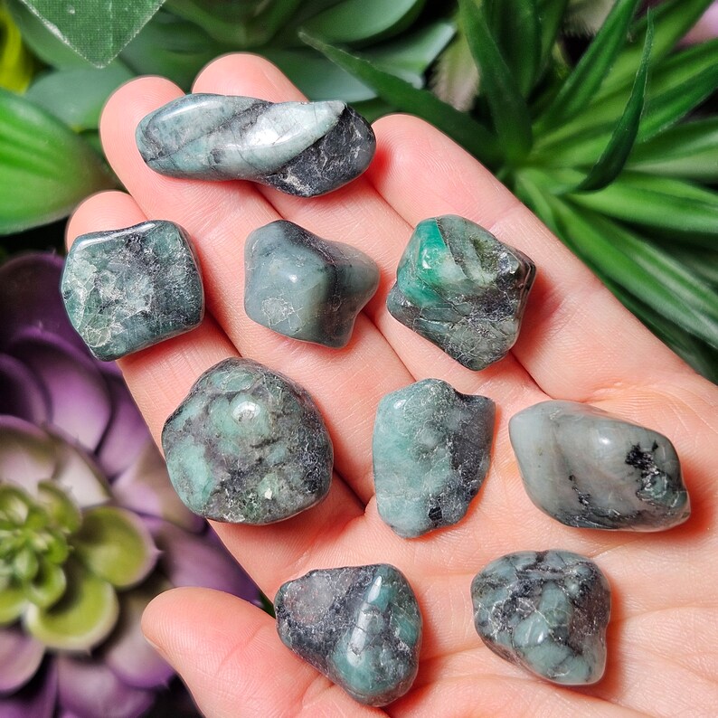 May include: A selection of emerald gemstones, each with a unique shape and varying shades of green and black. The stones are smooth and polished, showcasing natural patterns and textures. The gemstones are displayed in a hand.