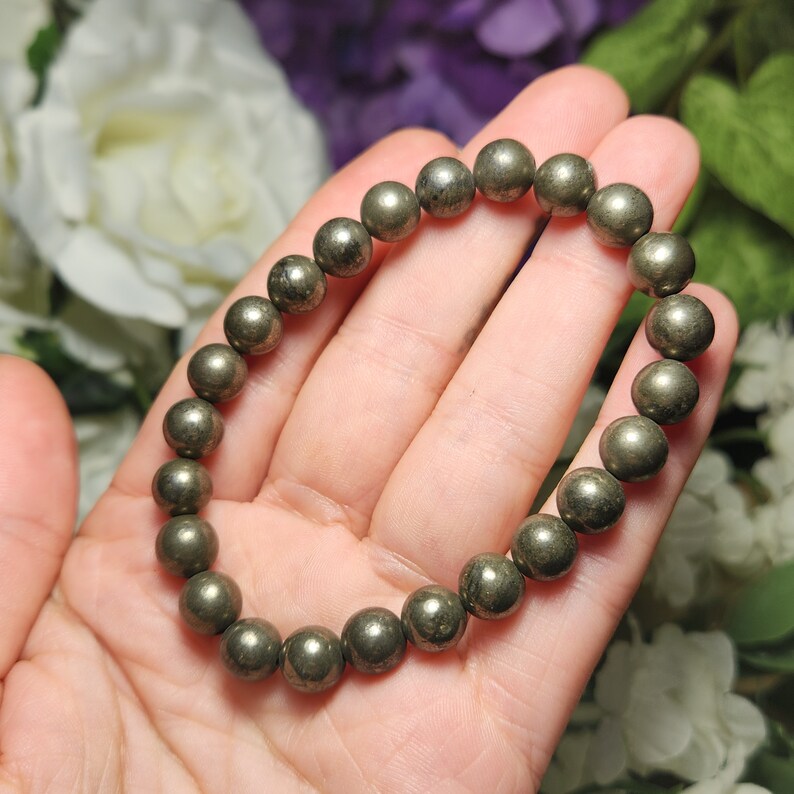 May include: A pyrite bracelet made of spherical, metallic beads. The beads are a muted gold colour and strung together to form a complete circle. The bracelet is held in a hand against a backdrop of white and purple flowers.