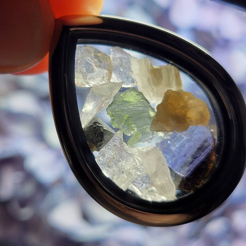 Puede incluir: Un colgante en forma de l&aacute;grima con un marco negro, lleno de varias piedras preciosas de colores y en bruto. Las piedras preciosas incluyen tonos claros, verdes, amarillos y azules, creando una exhibici&oacute;n vibrante. El colgante est&aacute; sostenido por los dedos de una persona.