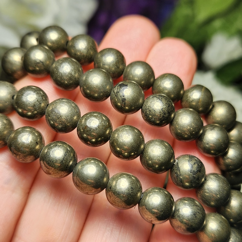 May include: A close-up of a pyrite bead bracelet. The beads are spherical, metallic, and have a golden-grey colour. The bracelet is held in a hand, with the beads in focus. The beads are approximately 1 cm in diameter.