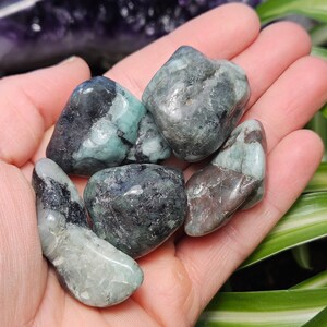 May include: A selection of tumbled emerald stones in shades of green and black. The gemstones are smooth and polished, displaying natural patterns. The stones are held in an open hand, highlighting their size and colour variations. Each stone is unique.