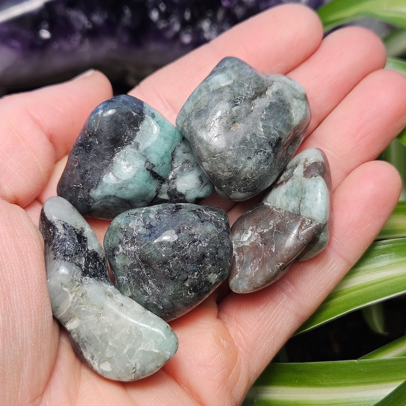 May include: A selection of tumbled emerald stones in shades of green and black. The gemstones are smooth and polished, displaying natural patterns. The stones are held in an open hand, highlighting their size and colour variations. Each stone is unique.