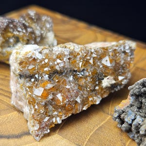 May include: Close-up of raw, uncut crystals with a mix of orange, brown, and white hues. The crystals have a rough, textured surface and are displayed on a wooden surface. The crystals are a natural mineral specimen.