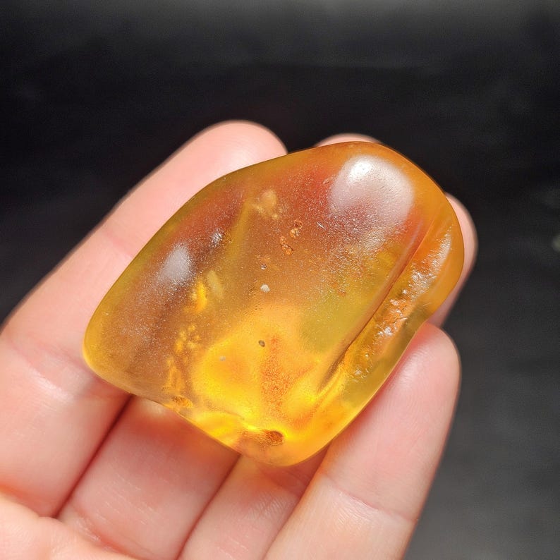 May include: A polished, translucent amber stone with a warm golden-orange hue. The organic, irregular shape features internal inclusions and natural variations in color density, creating a glowing effect when light passes through the smooth surface.
