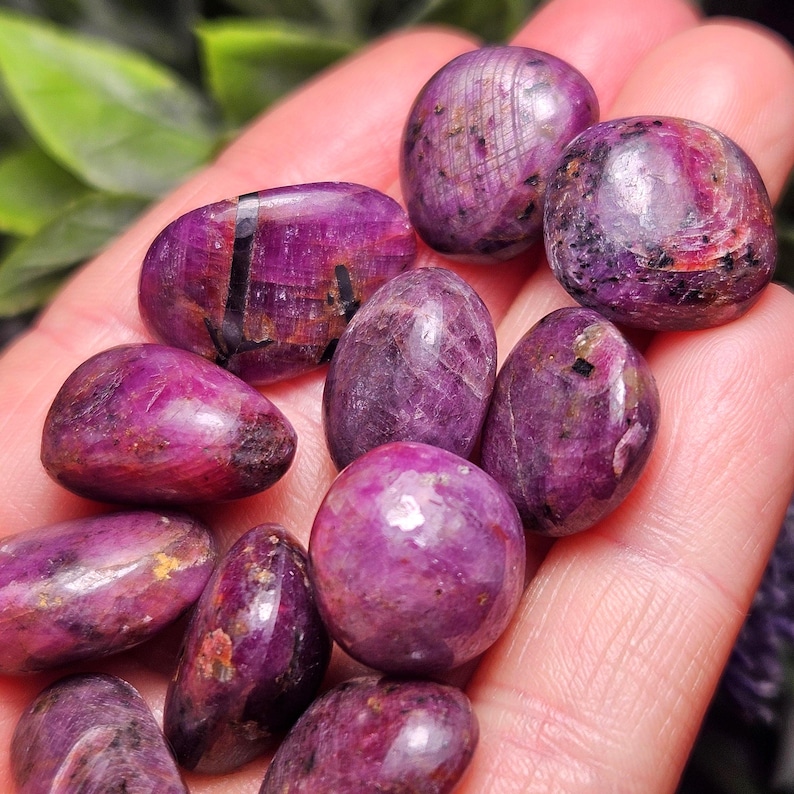 May include: A collection of polished, oval-shaped gemstones in varying sizes. The stones are a deep, rich magenta color with subtle variations in tone and texture. Some stones have dark inclusions. The gemstones are held in a hand.