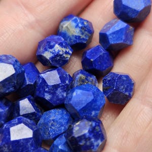 May include: A collection of small, faceted lapis lazuli beads. Each bead features a deep royal blue color with natural white calcite inclusions and subtle golden pyrite flecks. The stones are cut into multi-faceted shapes with smooth edges.