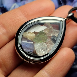 Puede incluir: Un colgante en forma de l&aacute;grima con un marco plateado, lleno de varias piedras preciosas de colores. El colgante cuelga de un cord&oacute;n negro. Las piedras preciosas incluyen cristales verdes, blancos, transparentes y azules.