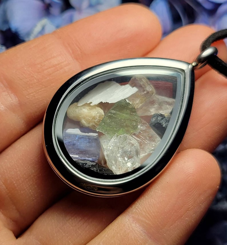 Puede incluir: Un colgante en forma de l&aacute;grima con un marco plateado, lleno de varias piedras preciosas de colores. El colgante cuelga de un cord&oacute;n negro. Las piedras preciosas incluyen cristales verdes, blancos, transparentes y azules.