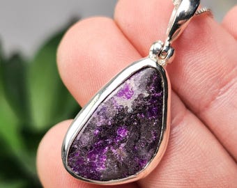 May include: A teardrop-shaped pendant featuring a textured, dark purple and black stone set in a polished silver-toned metal frame. The pendant includes a silver-toned bail at the top, designed to be worn on a chain or cord necklace.