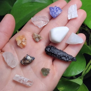 Puede incluir: Una colecci&oacute;n de varias piedras preciosas en tonos claros, blancos, naranjas, marrones, morados y negros. Las piedras preciosas son de diferentes formas y tama&ntilde;os, incluyendo un cristal claro y rectangular y una piedra blanca y redondeada.