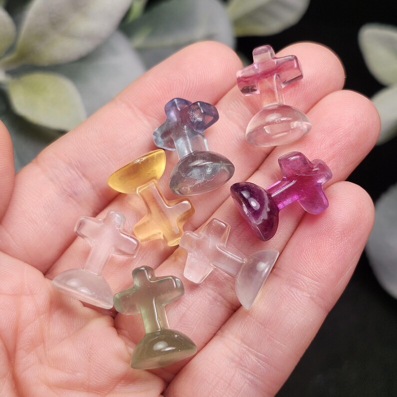 May include: A collection of small, colorful, cross-shaped crystal figurines. The crystals are in various colors, including clear, purple, yellow, and green. The figurines are held in a hand, with a blurred background of green leaves.