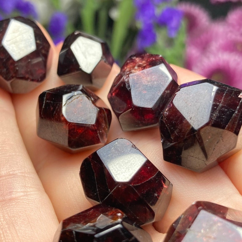 May include: A collection of eight red garnet crystals, each with a unique, faceted shape. The crystals are smooth and polished, with a deep red color.