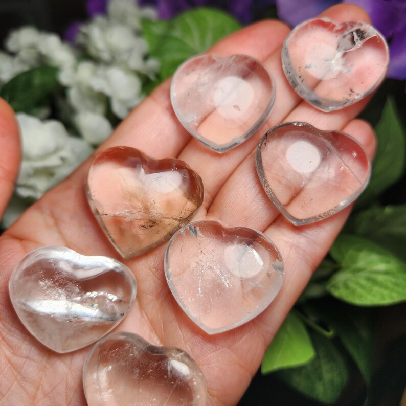 May include: Seven clear quartz heart-shaped crystals in various sizes, held in a hand. The crystals are translucent and have a slight shimmer.