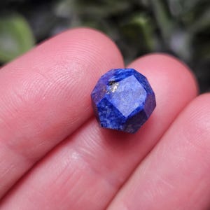 May include: A faceted lapis lazuli gemstone with a deep blue color and small golden pyrite inclusions. The stone is cut into a geometric, multi-sided shape with flat surfaces that reflect light, showcasing the natural mineral texture and deep blue hue.