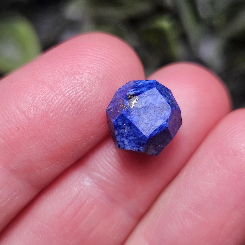 May include: A faceted lapis lazuli gemstone with a deep blue color and small golden pyrite inclusions. The stone is cut into a geometric, multi-sided shape with flat surfaces that reflect light, showcasing the natural mineral texture and deep blue hue.