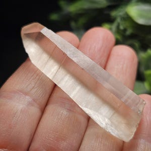May include: A clear, elongated quartz crystal point with a hexagonal shape and a pointed tip. The crystal features visible internal horizontal striations and a smooth, translucent surface that allows light to pass through its structure.