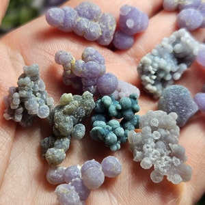 May include: A selection of small, rounded stones resembling grapes, in shades of lavender, grey, and teal. The stones are clustered, creating a textured look. The close-up image highlights the variety of colours and shapes of the stones.