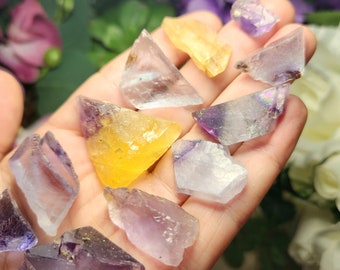 May include: A collection of small, faceted crystals in shades of purple, yellow, and clear. The crystals are arranged on a hand, showcasing their various shapes and sizes.