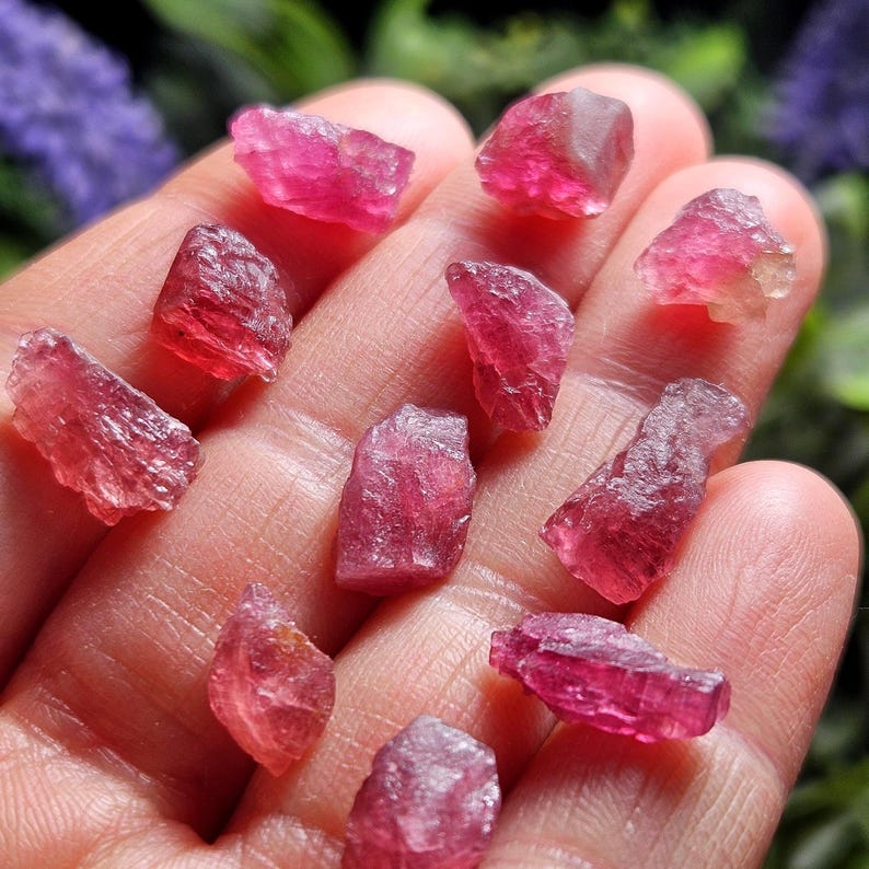 May include: A collection of small, raw, translucent pink tourmaline crystals with irregular, jagged edges and a vibrant magenta hue. These natural gemstone fragments exhibit a crystalline structure with varying levels of clarity and light reflection.