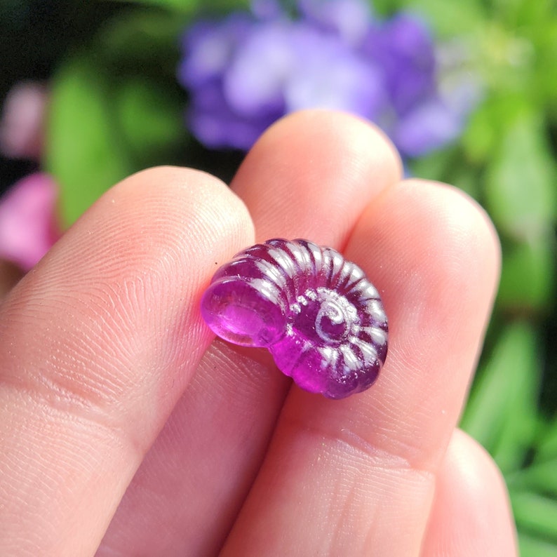 Peut inclure: Petit objet translucide en forme de coquillage violet. La coquille en spirale pr&eacute;sente des ar&ecirc;tes d&eacute;taill&eacute;es et une surface lisse et polie. L'objet est tenu dans une main, avec un arri&egrave;re-plan flou de feuillage vert et violet.