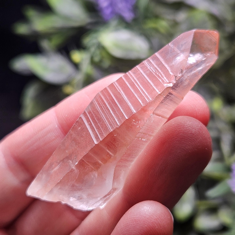 May include: A translucent, pale pinkish-clear quartz crystal point featuring distinct horizontal striations along its elongated, faceted surface. The crystal exhibits a sharp, pointed termination and a natural, irregular base with internal inclusions.
